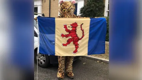 PETER JAMES BOWMAN A person dressed in a tiger mascot costume holds the Flag of the Fens, which has a red heraldic tiger facing left on a yellow background with a blue stripe on either side. The mascot is standing on a road in front of a white van and a house. 