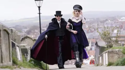 Danny Lawson/PA A man and a woman dressed in gothic clothing walk arm in arm through the graveyard near Whitby Abbey