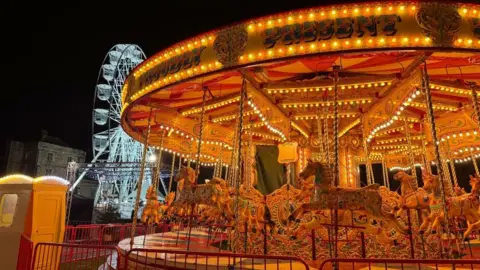 A picture of a merry-go-round lit with warm golden lights. In the foreground there is a ferris wheel with white lighting. There is a brown brick building behind that.