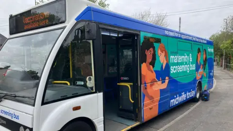 A single-decker bus parked on a roadside with maternity & screening bus printed on the side, with graphics of a nurse touching the stomach of a pregnant woman, and another of a woman holding a baby with a nurse looking on. The screen on the front of the bus says "vaccinations available here".