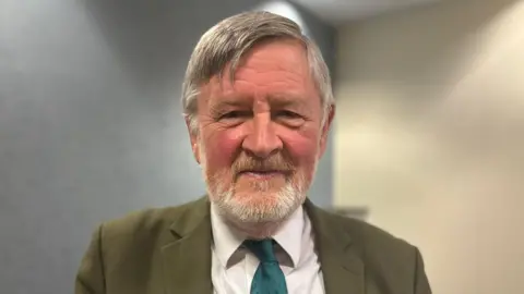 David Keating is standing indoors. He has grey hair and a beard and is wearing a green suit jacket over a white shirt and darker green tie.