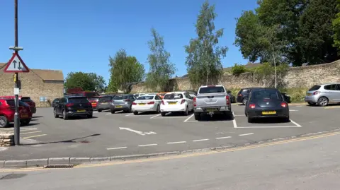 A view of a car park from a road on a sunny day - there are diagonal bays, mostly filled with cars, with an old wall down one side with trees.