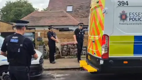 Three police officers wearing black short sleeved uniform and black and white police hats with chequers on, gather on a pavement near a large Essex Police van. It has neon yellow and orange chevrons on the back and is white and blue. Photo collages of dogs are propped up against a brick wall, with the headline "Still missing". There is a residential house behind the wall.
