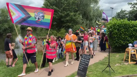 Two people at the front of the march hold a large banner with colourfully-dressed males and females walking behind them.