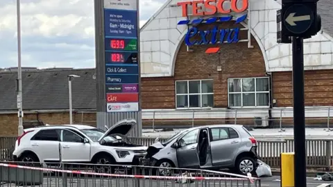 A Tesco Extra can be seen in the background of the shot along with the two collided vehicles before a metal fence. A police cordon is closing off the area.