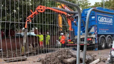 A digger lifts up a mound of earth as workers in fluorescent yellow and orange stand nearby with white safety helmets on
