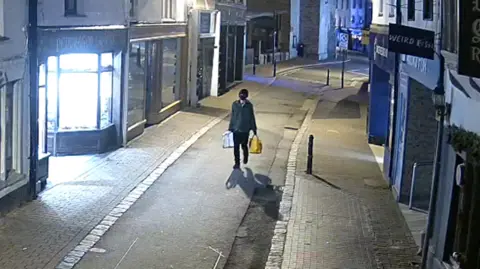 This is an image of a person with a black face covering. They are holding a large silver ladder and a yellow bag. They are walking on a stretch of street. It is dark.