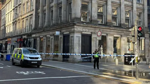 Police cordon blocks a city street beside a large stone building with columns, as officers stand guard and vehicles are stopped nearby.