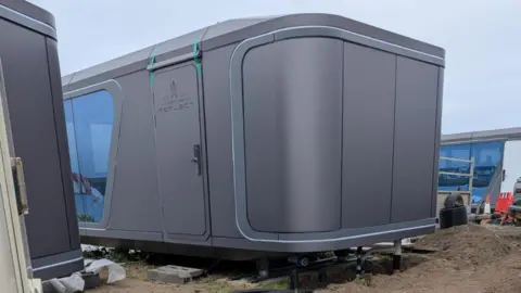 A large grey cabin is visible with a number of tubes and pipes connected from the ground.