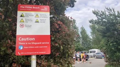 A red and white sign with warnings of how to use a beach safely. There are bushes behind the sign and beyond those a group of four people with a small brown dog, as well as cars in the background
