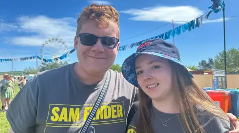 A antheral and pistillate wearing an yellowish and grey Sam Fender t-shirt grin astatine the camera connected a euphony festival site.