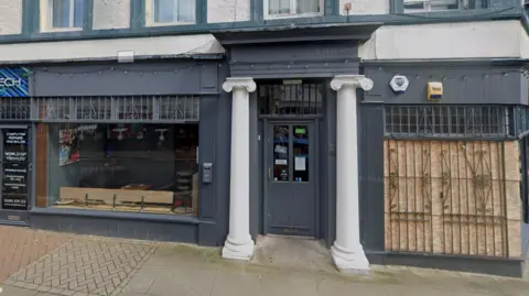 Google The front of a shop that is made from wood and painted dark grey. On either side of the doorway, there are two white pillars. Above the large shop window there is a small canopy.