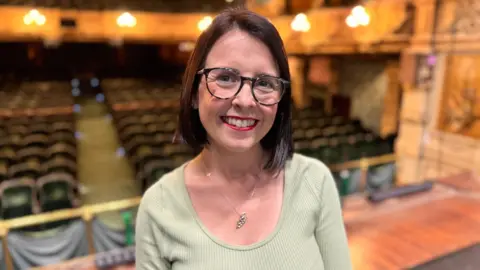 Sarah Maltby wears a green top and silver necklace that is in the shape of the Isle of Man. She also wears dark glasses, and red lipstick and has short dark hair. The view of the theatre from the stage can be seen behind her.