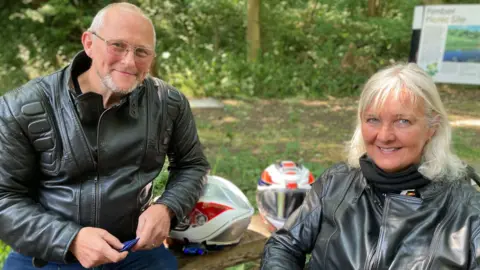Ivor is sitting on a picnic table on the left with grey hair and grey goatie, black biker's jacket and jeans and Karen is on the right, with shoulder-length white hair wearing a black jacket. Both are smiling at the camera
