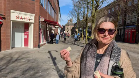 BBC Jane Kent en gafas de sol frente a la tienda de Gail comiendo un plátano