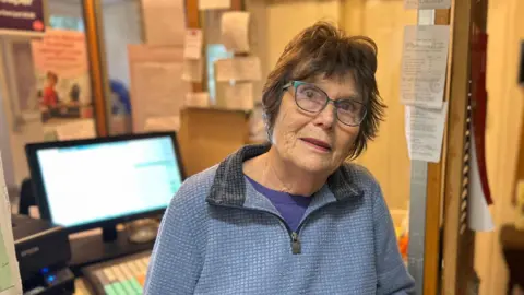 Jenifer Fell smiles at the camera inside her Post Office within her home. She has short brown hair and wears glasses along with a blue jumper. A computer can be seen behind her. There are many posters stuck on the walls around her.