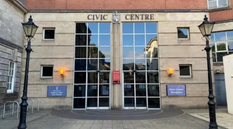 The Civic Centre in Stoke, a light-coloured brick building with large panelled windows which are about two-storeys in height. There are two traditional-style lampposts in the foreground.