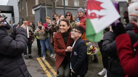 PA The Princess of Wales posing for a photo with a young boy with glasses wearing a black school blazer outside Hiut Denim. Crowds of people can be seen stood behind them, one person is holding a Welsh flag. The princess is also holding a bunch of tulips placed behind the boy's back while the photograph is being taken.