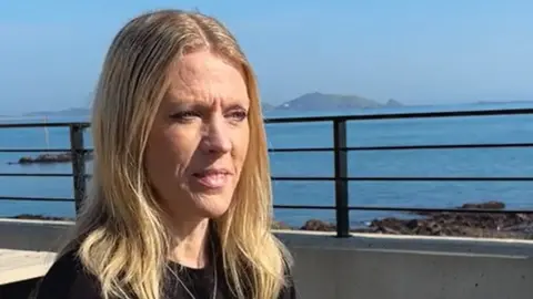 A woman with blonde hair sat in front of the sea, in a black top sitting on a bench 