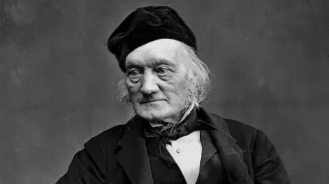 Getty Images A black-and-white portrait of a man with white long hair, wearing a velvet hat, black coat, waistcoat and bow tie and white shirt. He is gazing to the right and has a dark background behind him.