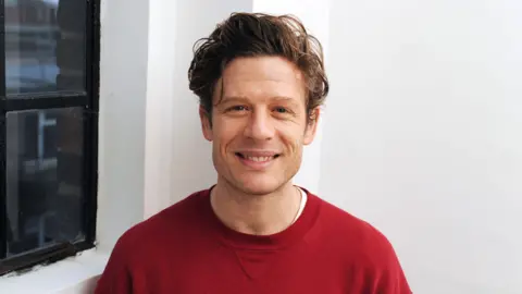 BBC James Norton looks at the camera, smiling. He is wearing a red jumper and standing in front of a white wall next to a black framed window.