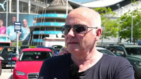 BBC A man with shaved white hair, looking slight off camera, he is in the car park of Manchester City stadium as the ground can be seen behind him. He has a black T-shirt and has sunglasses on. It is a head and shoulders shot.