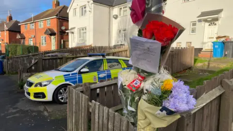 A police car parked up in front of a house with a number of flowers and tributes taped to a nearby lamppost