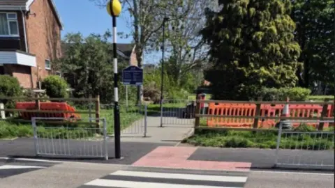A pedestrian crossing leading to a park with a blue sign at the entrance