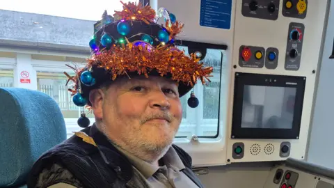 A man sat in the driver's seat in a train cab looking at the camera. He is wearing a festive hat with orange tinsel and small blue baubles on it.
