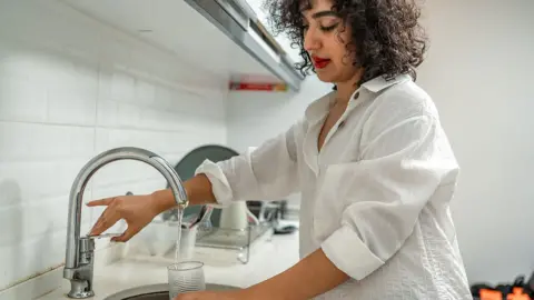 A woman fills a glass with water from a kitchen tap at home, she's wearing a white shirt and the kitchen has white tiles and a dish drying rack just visible behind the sink.