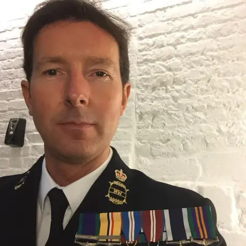 Terry Stewart A clean shaven man with short brown hair and dark eyes looks at the camera. He wears a Royal Navy blazer, white shirt and dark tie. On his chest are six medals hung on colourful ribbons. 