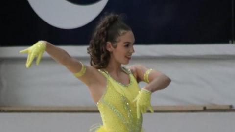 A girl with brown hair in a yellow dress and yellow gloves dancing