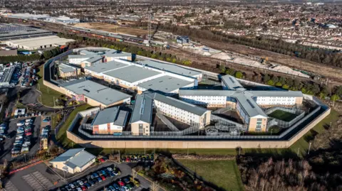 Aerial image showing prison in blocks and square flat topped roof buildings surrounded by numerous car parks and a distant railway line