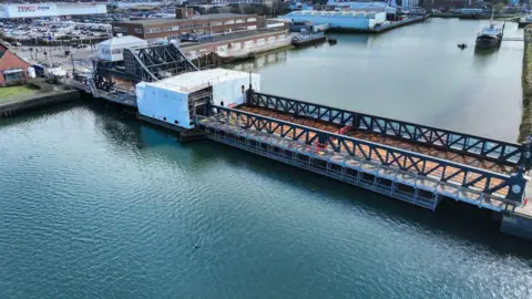 A bird's eye view of a bridge across a river. It has black sides. There is protective sheeting over part of the bridge where restoration  work is taking place. A boat is further down the river and there is a Tesco Extra on shore in the distance.