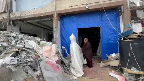 Sanaa stands outside her makeshift shop, holding part of a white wedding dress on a mannequin which stands in front of a big pile of rubbish and rubble