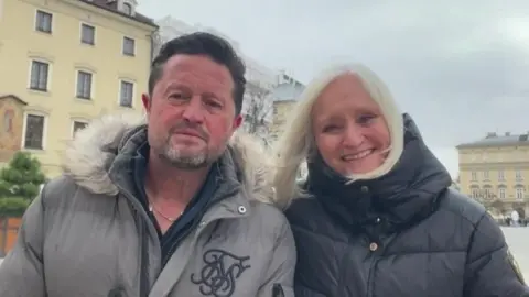 Mr Fenton with short dark hair wearing a grey anorak with white hood standing next to Mrs Fenton, who has long blonde hair, wearing a dark-coloured anorak. She is smiling. They are in a square in Poland - a historical building is visible in the background.