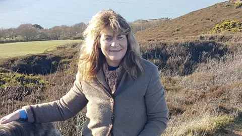 Annabel Harrison Annabel Harrison stands on a hill with a view of the sea in the background. She has wavy, fair hair and wears a brown jacket and patterned scarf.