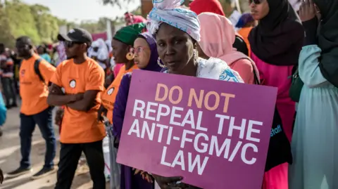 AFP An anti Female Genital Mutilation (FGM) protester holds a placard outside the National Assembly in Banjul on march 18, 2024, during the debate between lawmakers on a highly controversial bill seeking to lift the ban on FGM. Gambian lawmakers voted on March 18, 2024 to advance to the next parliamentary stage a highly controversial bill that seeks to lift a ban on female genital mutilation (FGM), which has been in place since 2015. The issue has divided the tiny West African nation for months, with hundreds gathering to protest outside parliament. (Photo by MUHAMADOU BITTAYE / AFP) (Photo by MUHAMADOU BITTAYE/AFP via Getty Images)