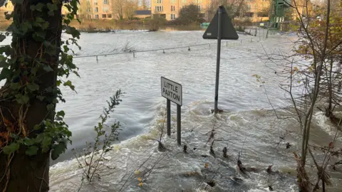 Harriet Heywood/BBC A road is submerged in water, visible above the water is a sign for Little Paxton. In the distance are flats.