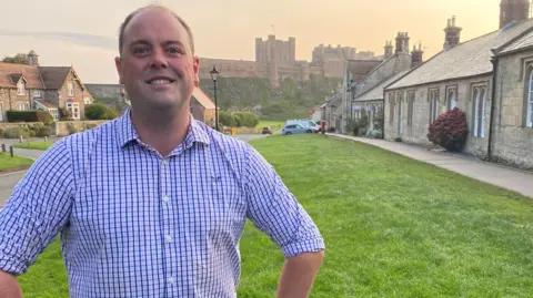 Guy Renner-Thompson is standing in front of Bamburgh Castle wearing a blue shirt and with his hands on his hips. 