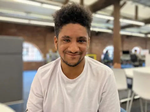A young man in a white t-shirt smiles at the camera. The background is blurred behind him but a table and chairs are visible.