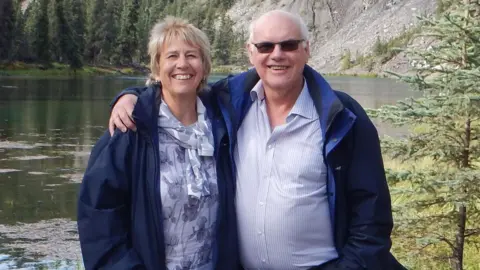 Mrs Barlow and Mr Greenaway stand in front of a lake, surrounded by pine trees. The couple smiles at the camera. Mr Greenaway has his arm around his wife's shoulder.