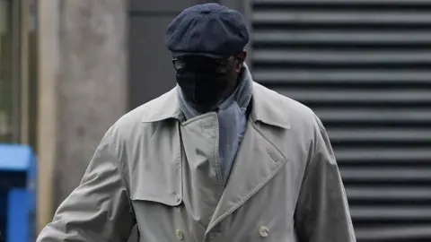 Charles Ehikioya wearing a beige trench coat, grey scarf, glasses, a black mask covering his face and a grey hat.