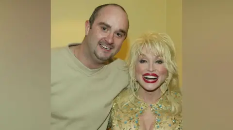 Supplied Joe Skelly has his arm around Dolly Parton. They are both smiling brightly at the camera. Skelly has very short, dark hair and is wearing a green t-shirt. Dolly has long blonde hair and is wearing bright red lipstick and a bedazzled yellow top.