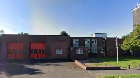 Google Maps A street view image of the fire station, a red brick, two-storey building. On the left two sets of tall, red garage doors allow access for appliances.