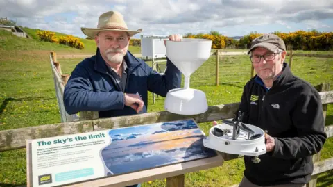 Paul is wearing a wide brimmed hat and holding what looks like a large white plastic chalice. Donald is wearing a peaked cap and holds a white base that houses a some tubes and machinery.