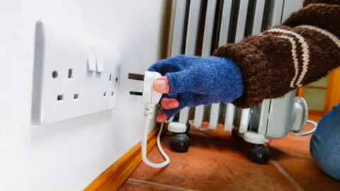 Getty Images A woman wearing a blue fingerless glove and a brown woollen jumper plugs a portable electric heater into a mains socket.