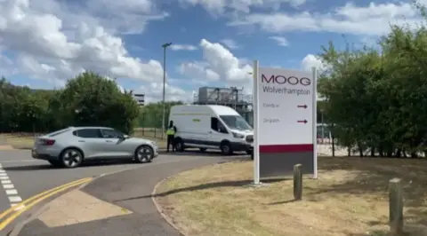 A silver car and a white van at the entrance to a site with metal fences and a white sign with Moog in red lettering on it
