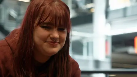 Kady is smiling looking down at a console. She's wearing a burgundy hoodie and has dyed red hair which is down to her shoulders. She has a nose ring and lip piercings.