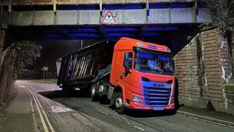 West Mercia Police A large lorry with an orange cab and a dark blue trailer is wedged underneath a railway bridge. 